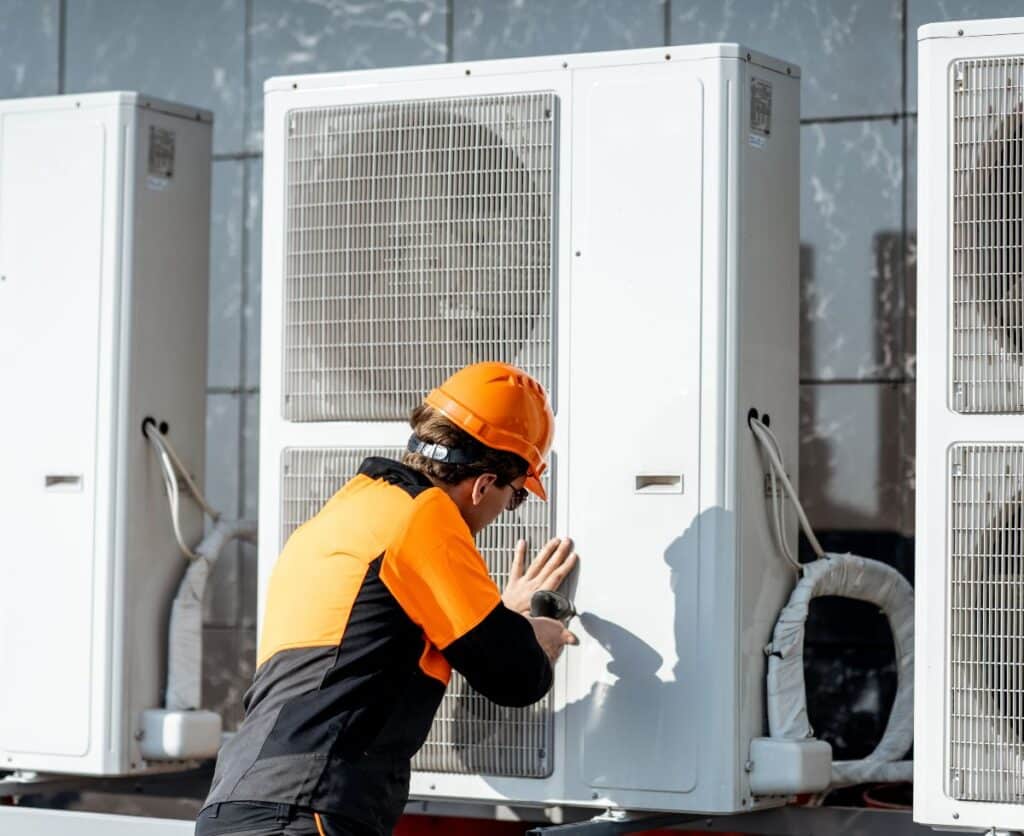 A man in an orange hard hat and safety vest inspecting a large heat pump unit.